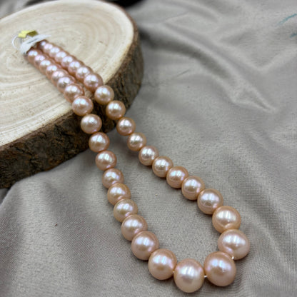 A double-strand pearl necklace with large, lustrous light pinkish-beige pearls, draped over a round wood slice on a draped taupe fabric background.