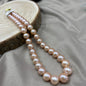 A double-strand pearl necklace with large, lustrous light pinkish-beige pearls, draped over a round wood slice on a draped taupe fabric background.