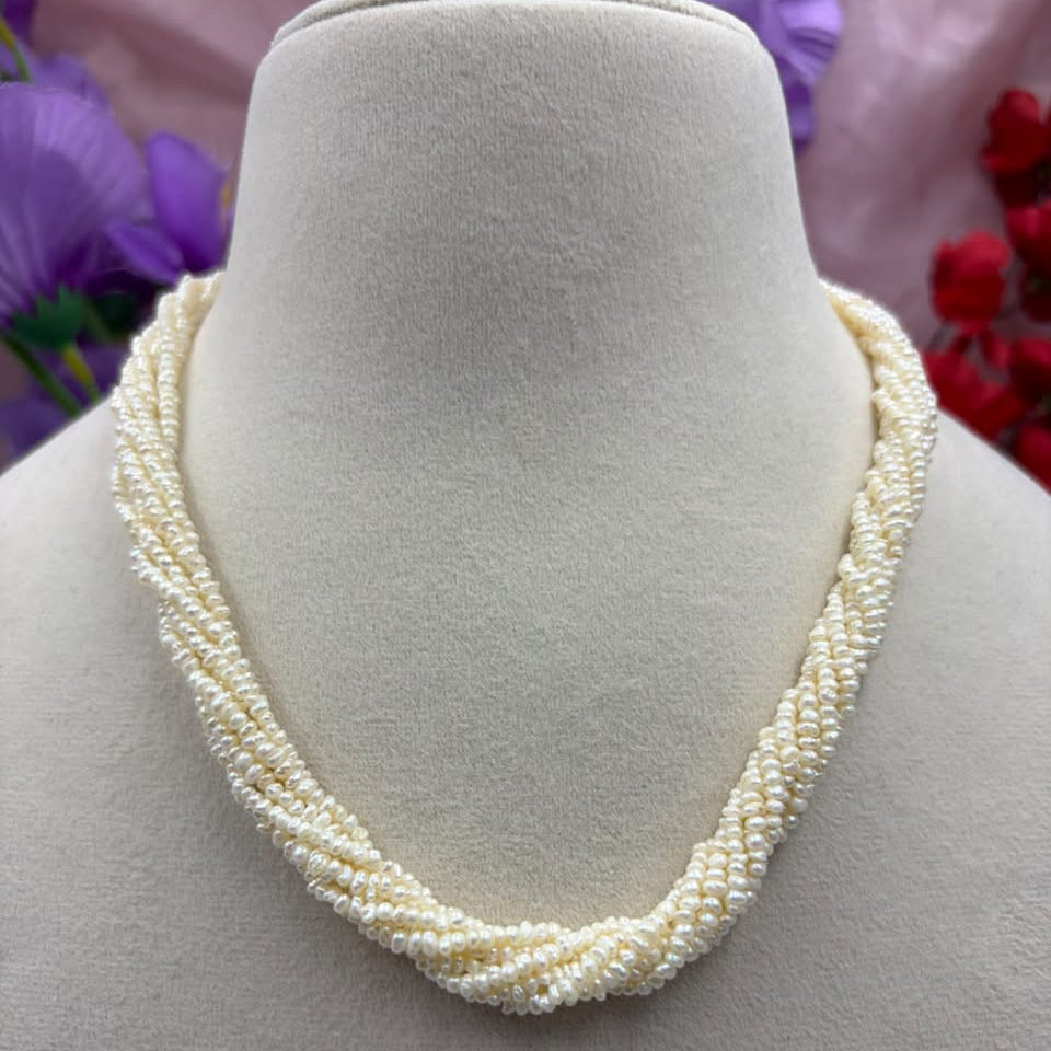 A multi-strand cream-colored pearl necklace displayed on a beige fabric jewelry bust, with blurred purple and red flowers in the background.