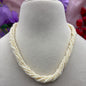 A multi-strand cream-colored pearl necklace displayed on a beige fabric jewelry bust, with blurred purple and red flowers in the background.